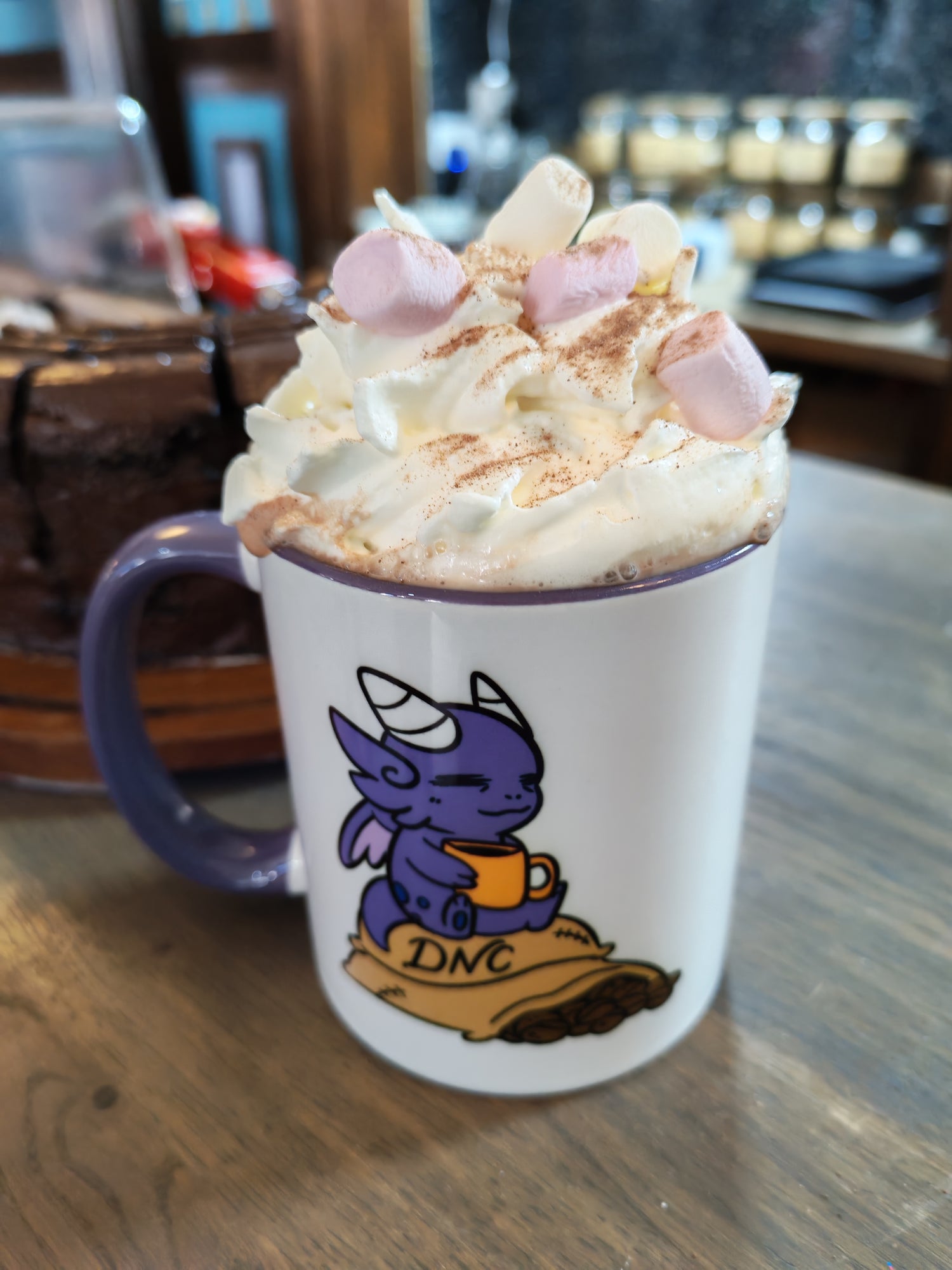 Mug topped with whipped cream and marshmallows. Mug also shows moki character holding a coffee cup